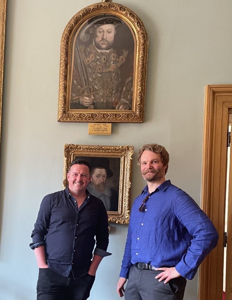 Two men stand in front of a wall displaying framed portraits. The upper portrait is of Henry VIII in regal attire, holding a sword. Below it is another framed portrait of a different historical figure. The man on the left wears a dark shirt and has short, dark hair, while the man on the right wears a blue shirt and has light hair and a beard. Both men are smiling and standing with their hands casually in their pockets or on their hips. The setting appears to be an art gallery or museum.