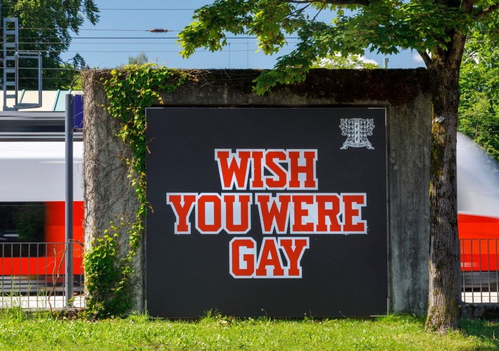 a large poster on a slab of stone in a public urban setting, it is black and has red letters saying 'wish you were gay'