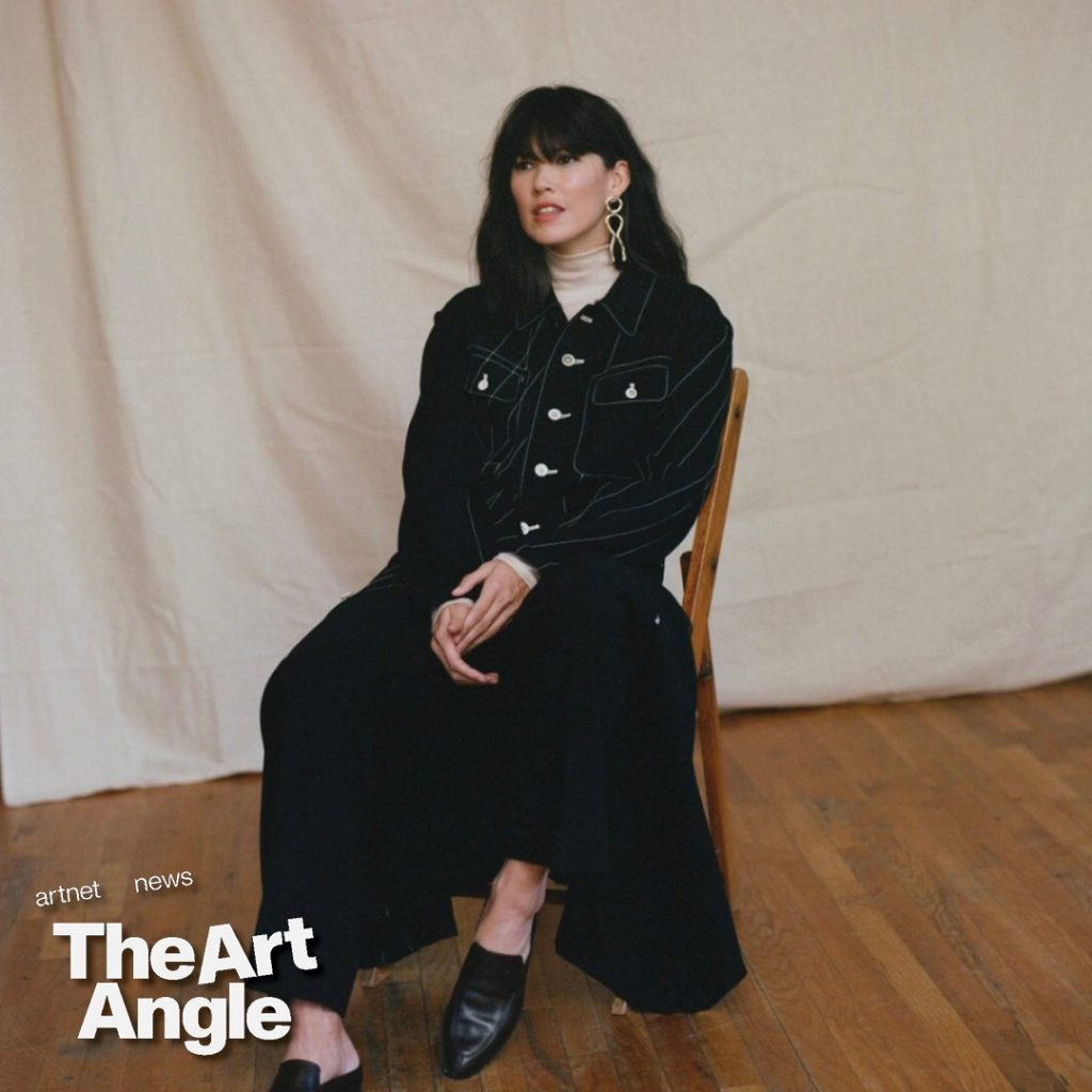 a white woman dressed in all black sits on a wooden chair against a white sheet backdrop