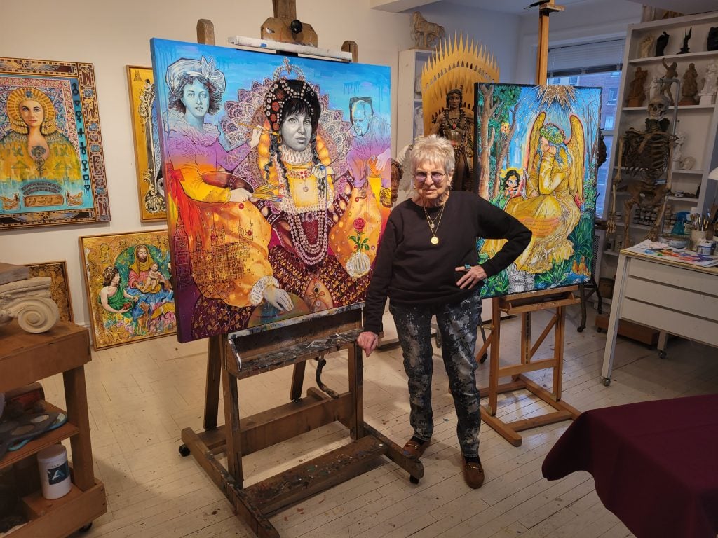 The elderly Audrey Flack, with short gray hair, stands in her art studio with her colorful recent paintings, two displayed on easels and others hanging on and propped up against the wall of her art studio.