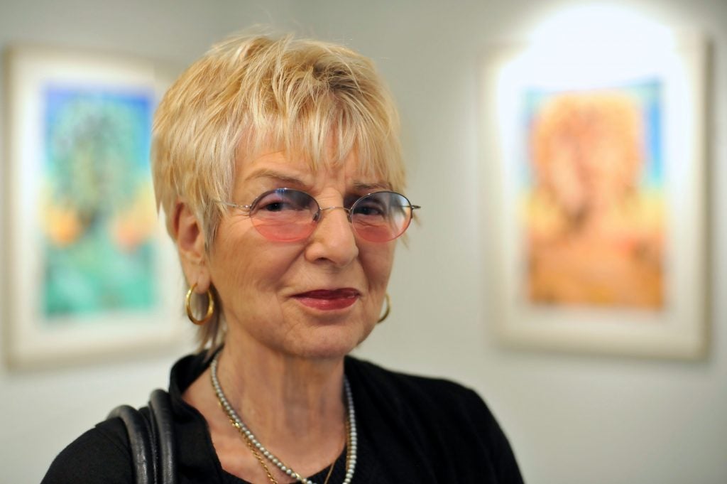 A photograph of artist Audrey Flack as an elderly woman wearing glasses, seen from the shoulders up, with two of her paintings in an art gallery behind her.