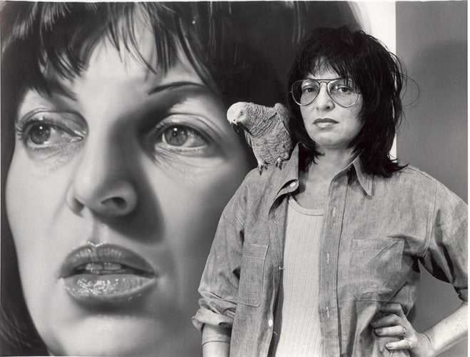 A black and white photograph of painter Audrey Flack, who died at 93, as a young woman. She has dark hair and glasses, and a parrot on her shoulder. She is standing in front of a larger than life self portrait of her face.