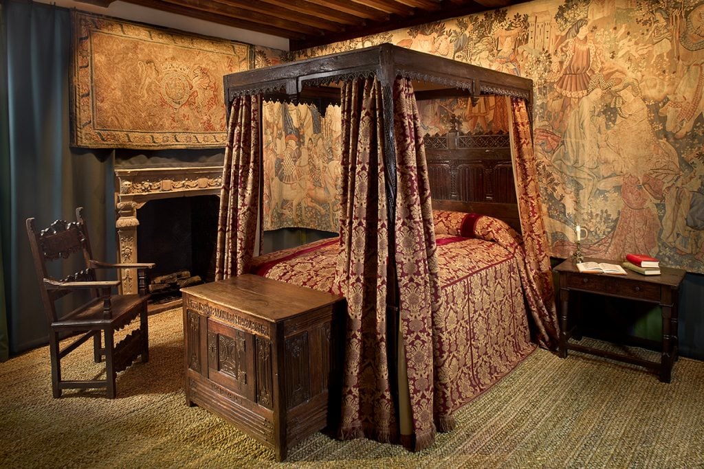 a large old fashioned interior with a four poster bed and tapestries on the wall and bits of old wooden furnitures