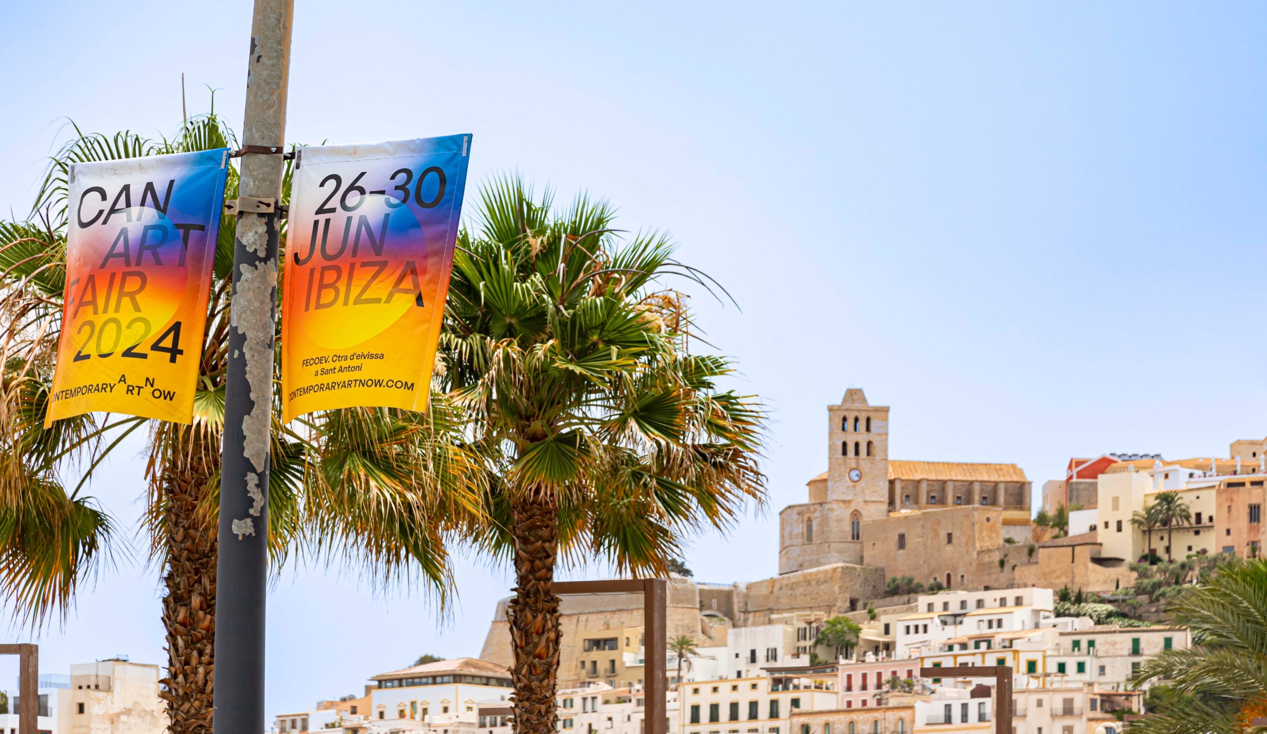 a colorful sign reading "26-30 JUN IBIZA" hands on a street light in front of palm trees, a blue sky, and attractive white buildings behind