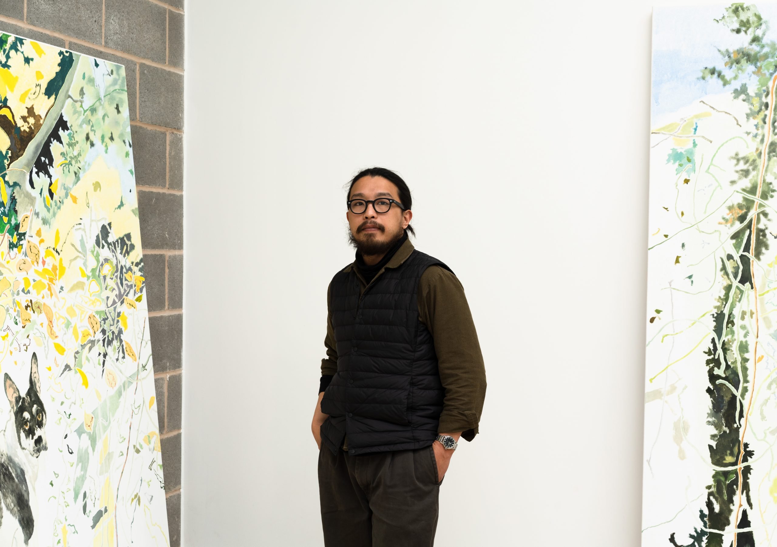 An East Asia man in black and glasses standing in between two large paintings.