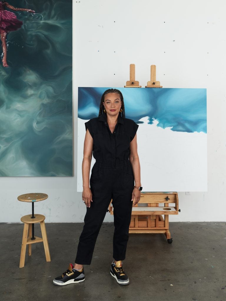 a black woman wearing a black top, pants, and sneakers stands in front of a half-finished painting 