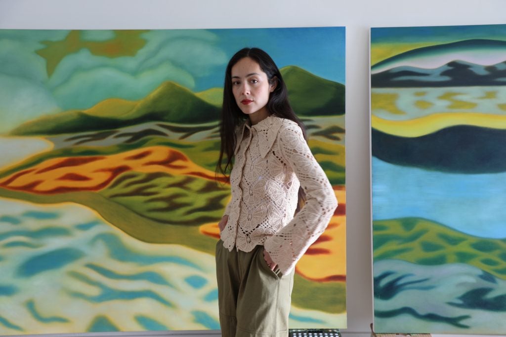A woman in her thirties with long dark hair stands in front of two large-scale landscape paintings.