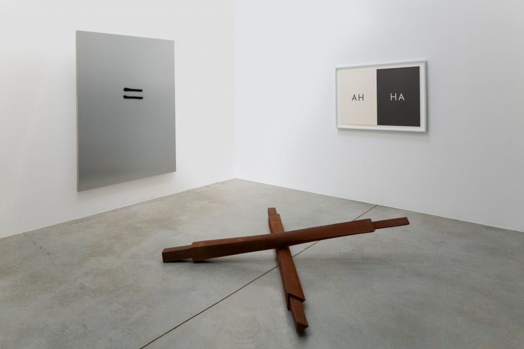 a white interior space that is brightly lit with a simple floor sculpture in the middle and then a silver surface with two small black horizontal lines in the middle and another work with the work "AH" on white and then "HA" on black