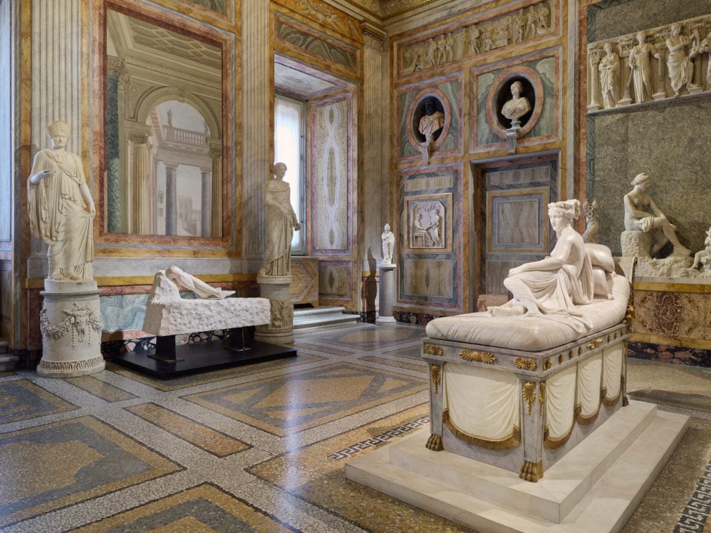 a large very ornate and grand old fashioned interior filled with marble statues but one is in a glass box and is clearly modern, it is two calves and feet protruding from a slab of rock