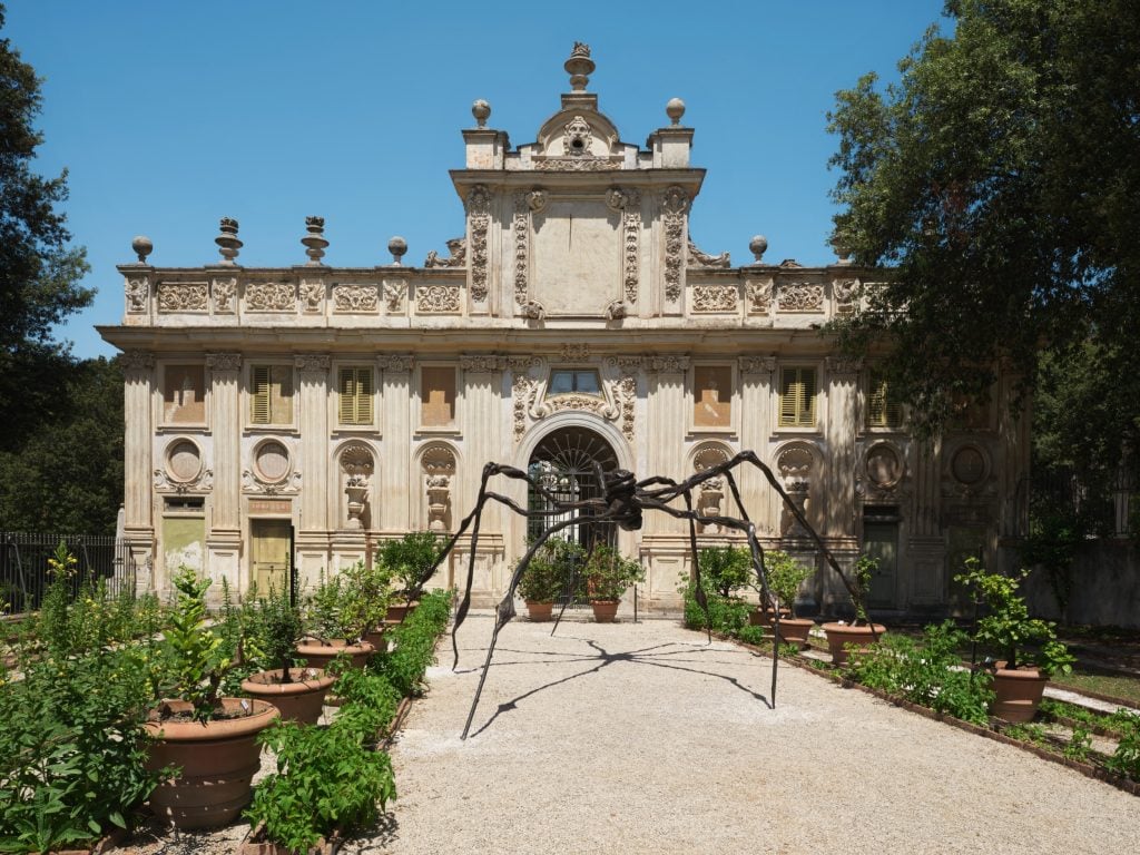 a large metal spider with spindly long legs installed outside an old fashioned but large and grand building such that you would need to walk under it in order to get in (as it takes up most of the path)