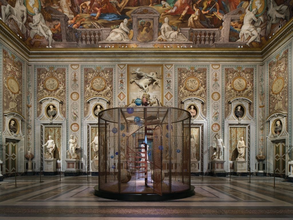 a circular cage filled with little contemporary sculptures sits in the middle of a very grand and ornate old fashioned interior 