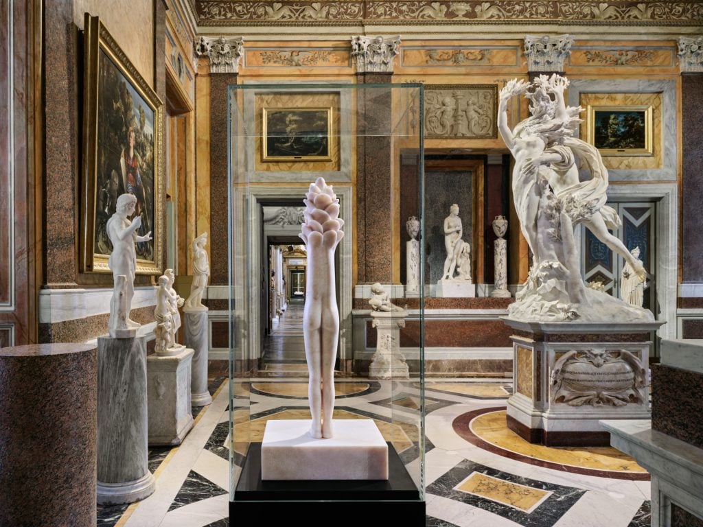 a large very ornate and grand old fashioned interior filled with marble statues but one is in a glass box and is clearly modern, it looks a bit like a long female figure