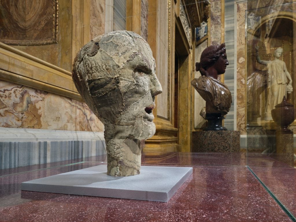 a large very ornate and grand old fashioned interior filled with marble statues but one is in a glass box and is clearly modern, it is near old stone busts but it is made of murky fabric 