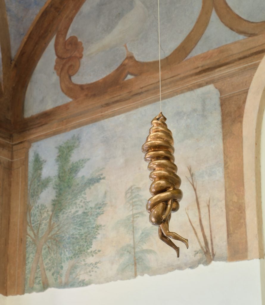 a strange bronze sculpture that is a bit like a cocoon with legs sticking out of the bottom hangs from a string in front of an old fashioned grand fresco painted on a wall