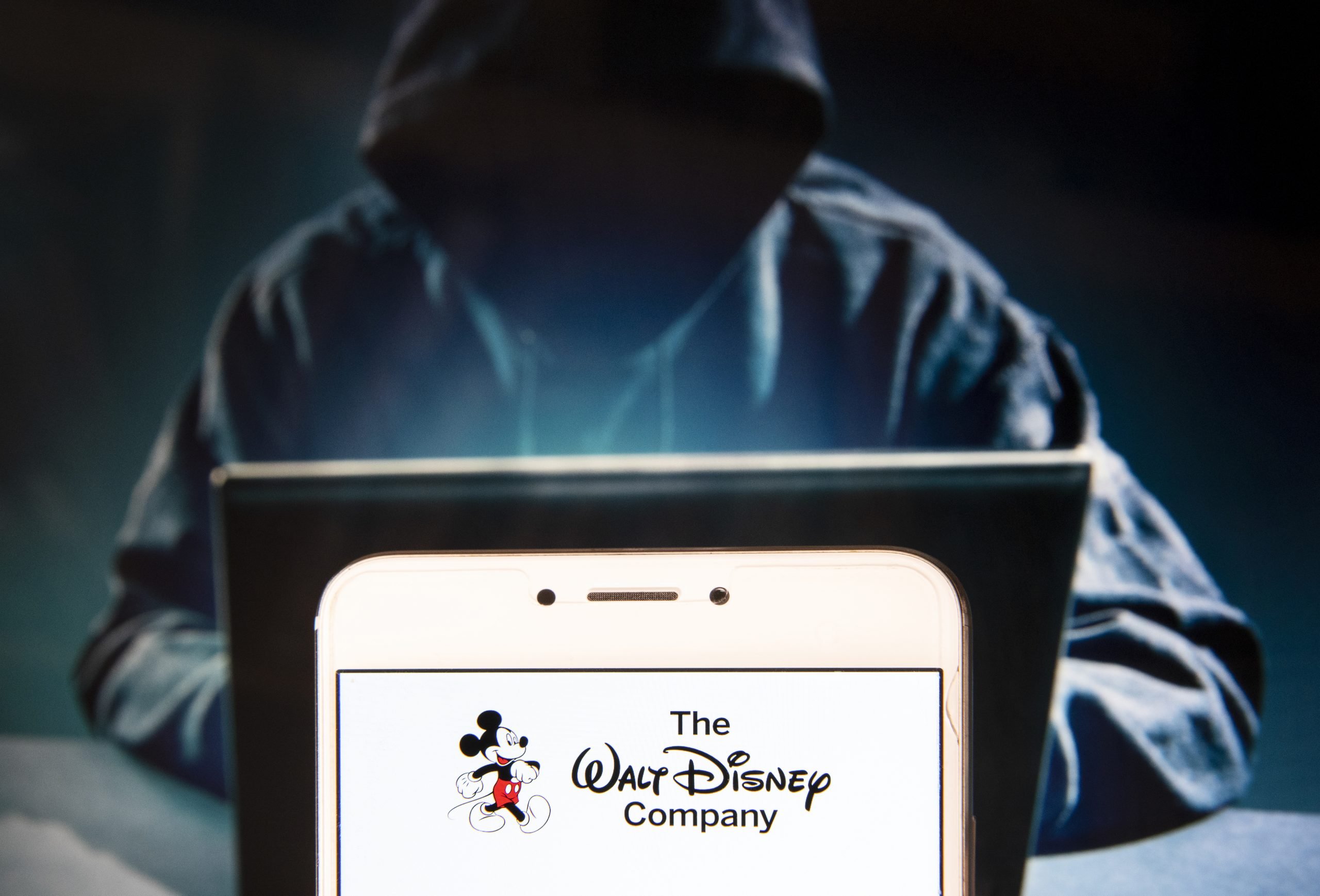 a hooded figure with no visible face is seen typing on a laptop, in the foreground is a phone screen with "the Walt Disney Company" text and Mickey Mouse logo on it