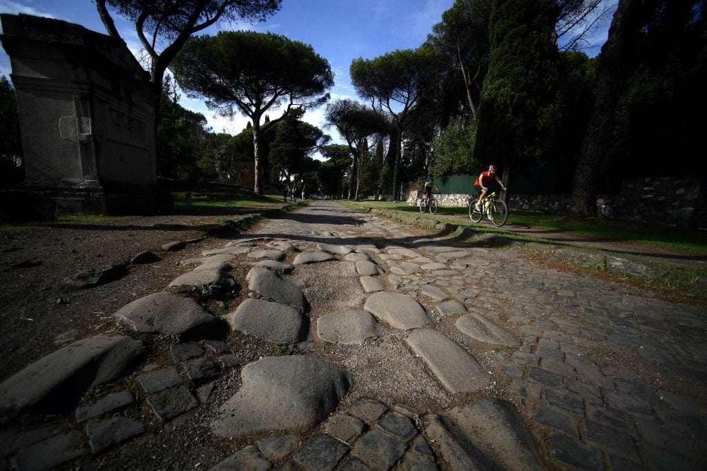 a cyclist rides down the Appian Way in Rome. There is a stone covering and large umbrella pines in the background 
