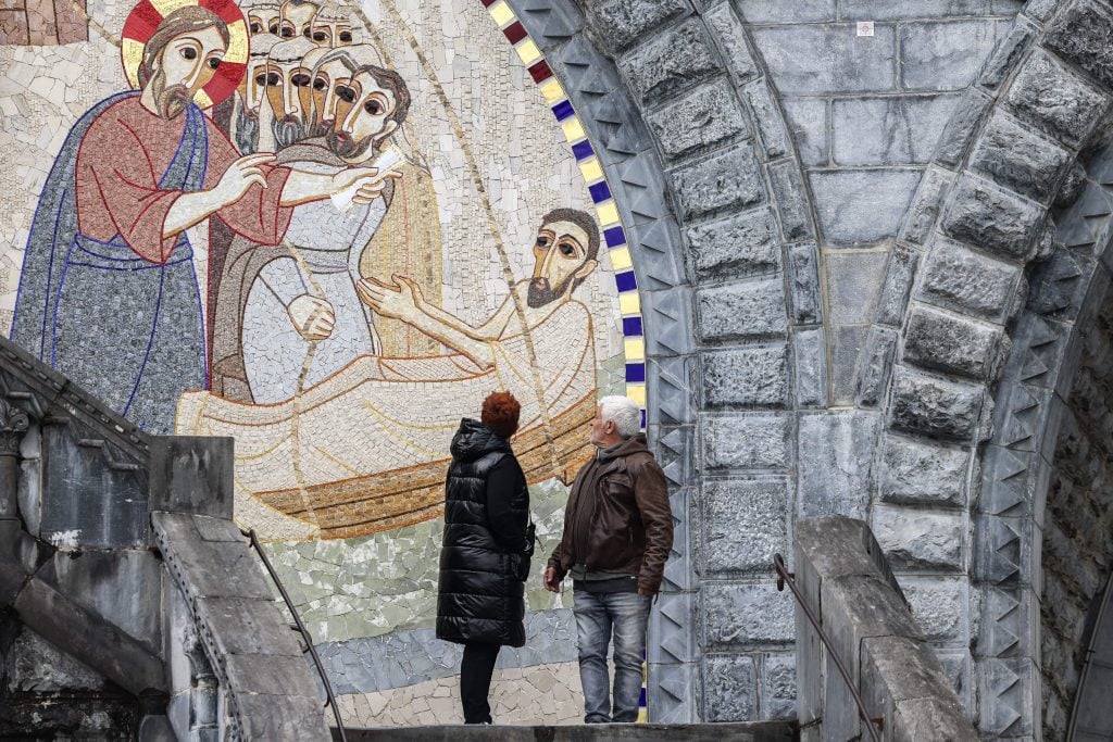 A man and woman stand on stone steps, admiring a large, detailed mosaic mural depicting a biblical scene with Jesus and several disciples. The mural is set into a stone archway, with intricate brickwork framing the artwork. The woman is dressed in a black coat, while the man wears a brown jacket and jeans. The scene captures their reverence and interest in the religious artwork.