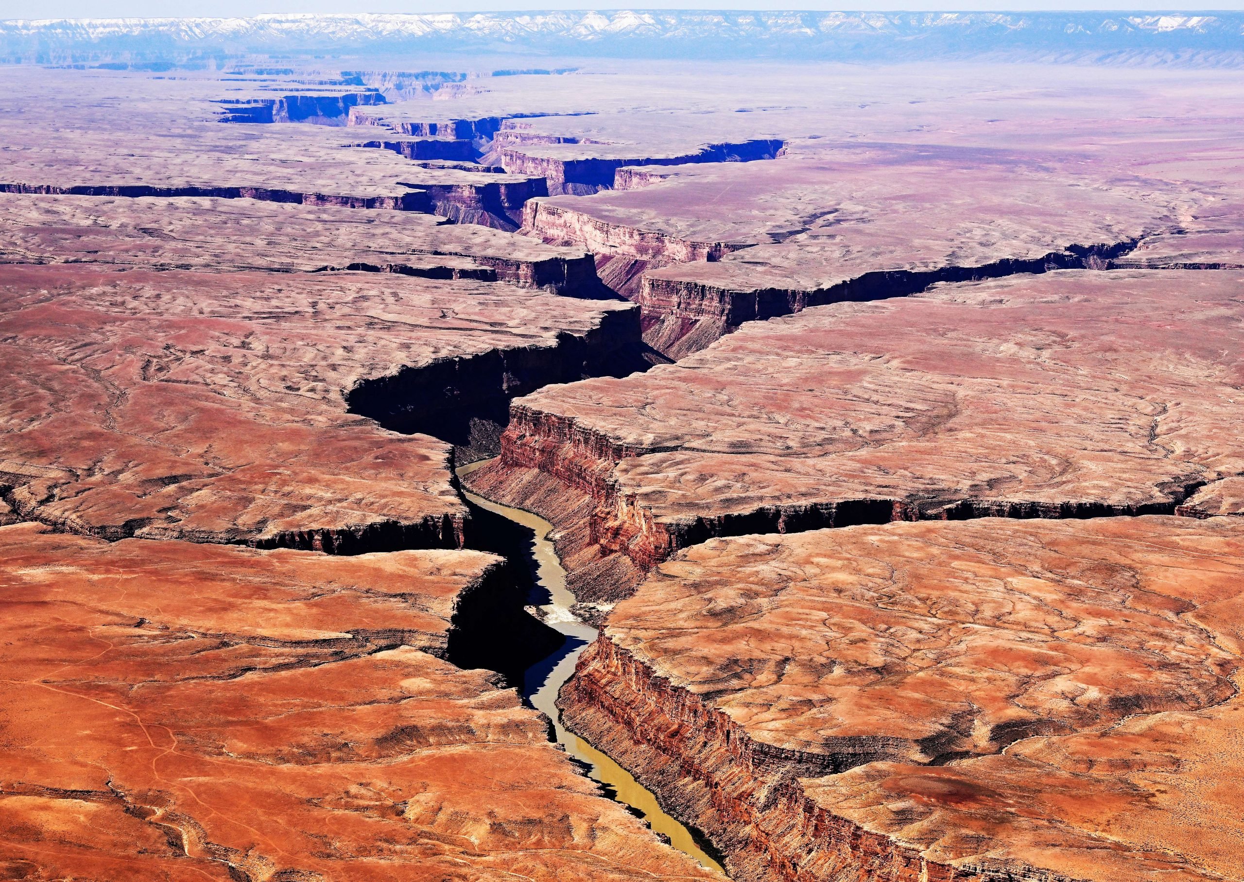 Aerial view of a vast desert landscape featuring a deep, winding canyon with steep cliffs and a river flowing through its base. The terrain is rugged and arid, with sparse vegetation and reddish-brown earth, extending into the horizon where snow-capped mountains are visible in the distance.