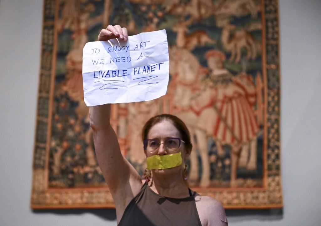 a protestor holds up a sign in front of a medieval tapestry at the Met Museum