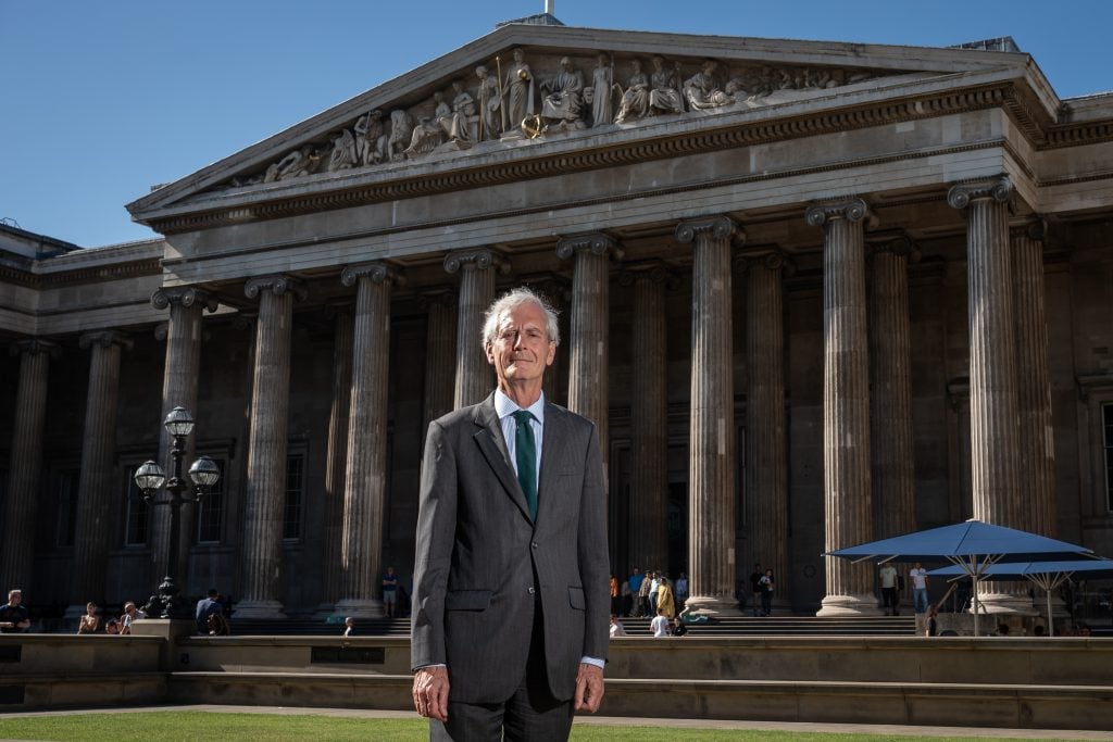 a suited elderly man stands outside a classical style building that is the British Museum, the sky is blue