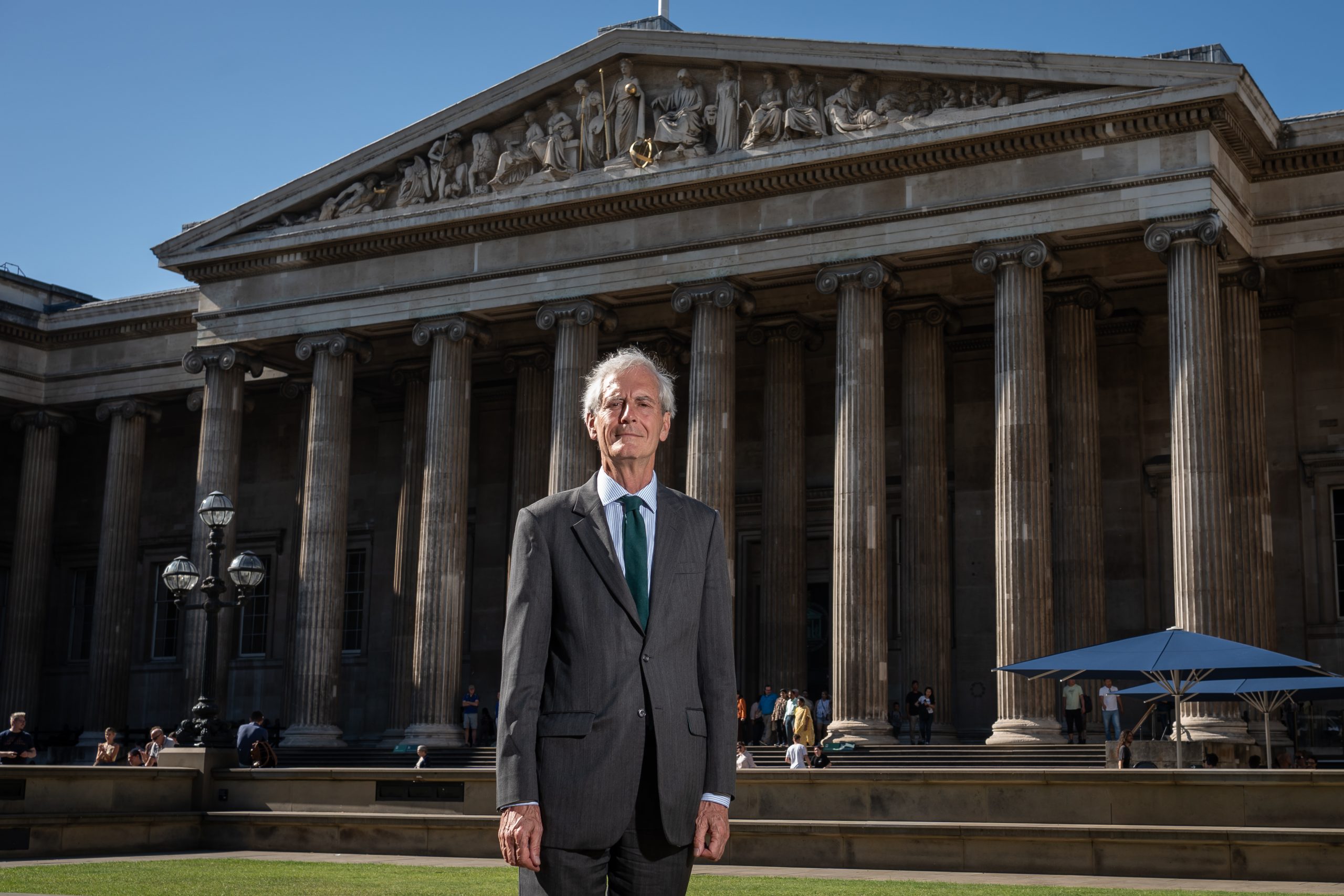 a suited elderly man stands outside a classical style building that is the British Museum, the sky is blue