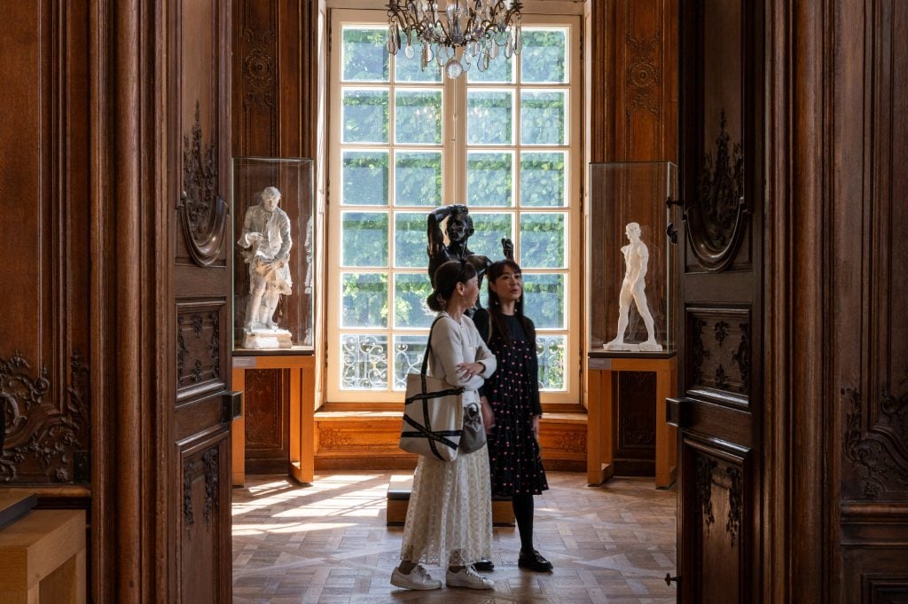 two women walk through a wood-panelled art gallery with sculptures