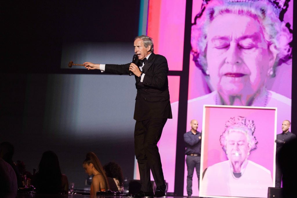 An auctioneer in front of a blown up screen showing a pink portrait of queen elizabeth ii