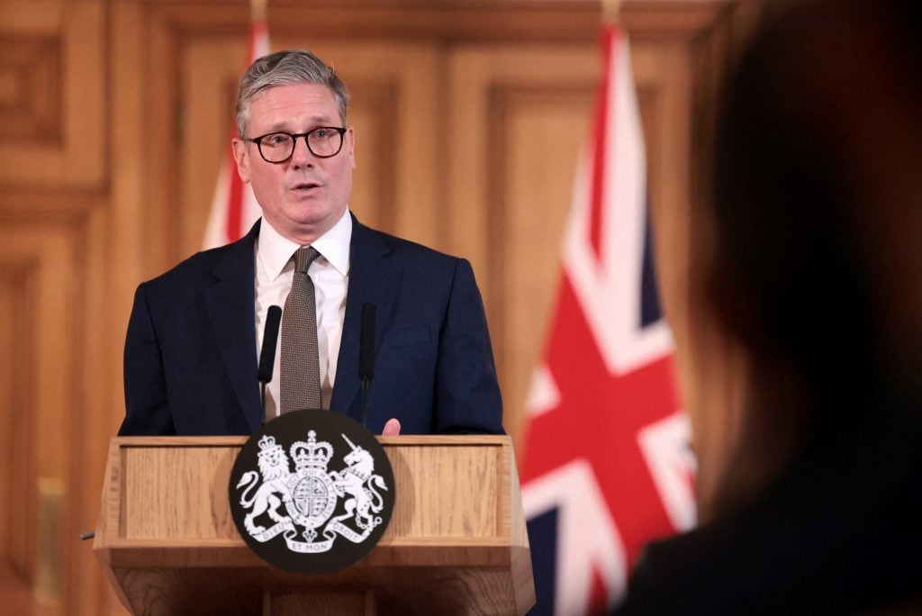 a man in a suit speaks at a podium with the UK flag behind him