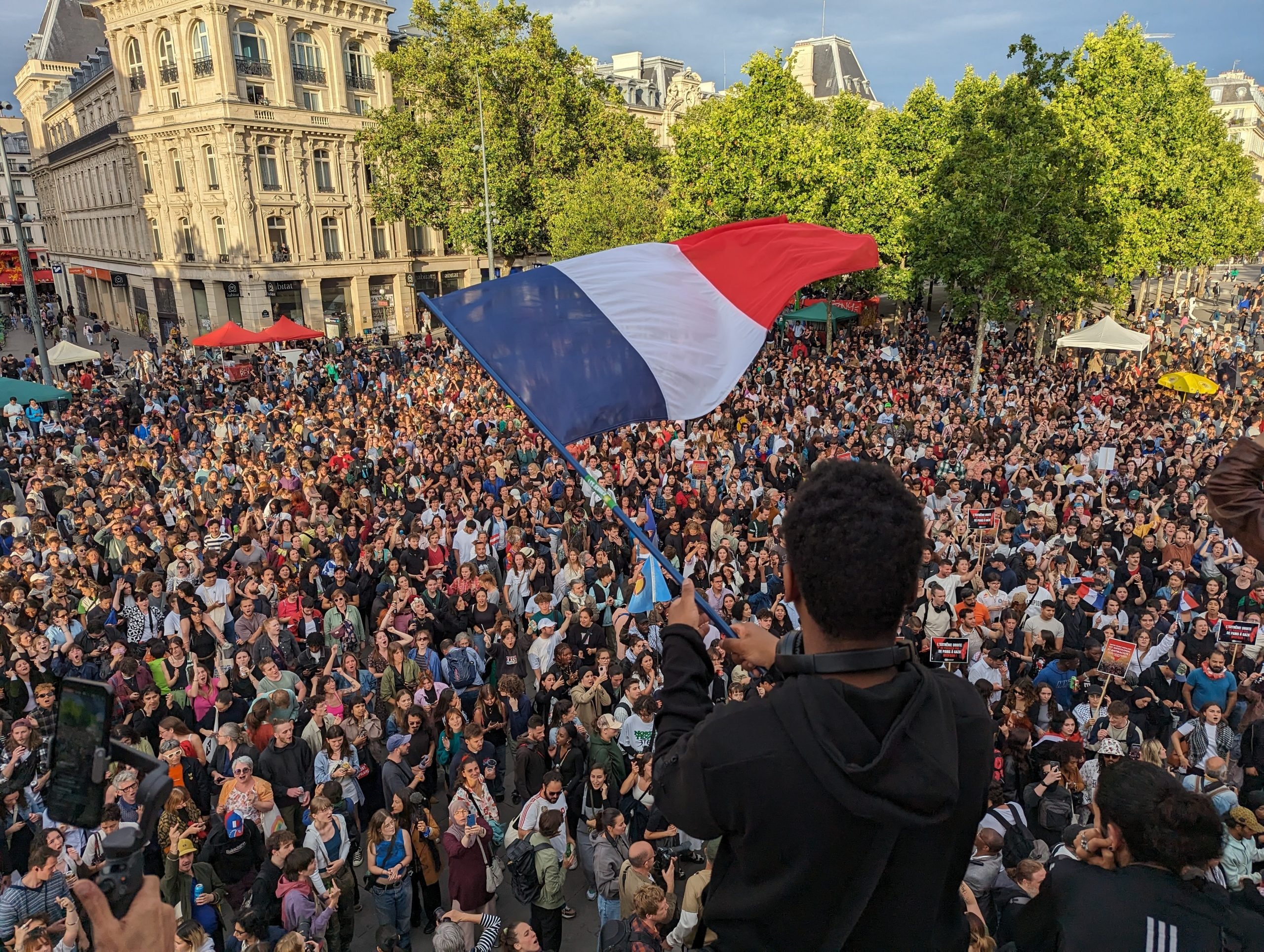 a person waves a french flag above a large crowd of people gathered in the street below