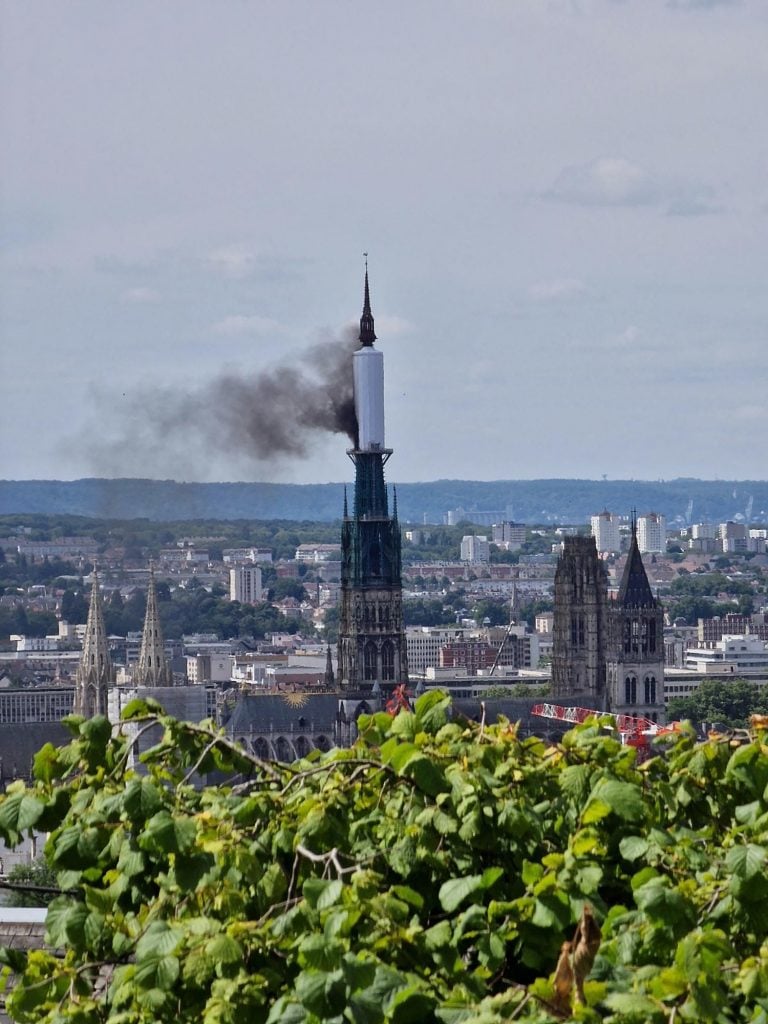 A cathedral tower with a black plume of smoke emerging from its spire against a backdrop of a cityscape and distant hills. The foreground features green foliage partially obscuring the view.