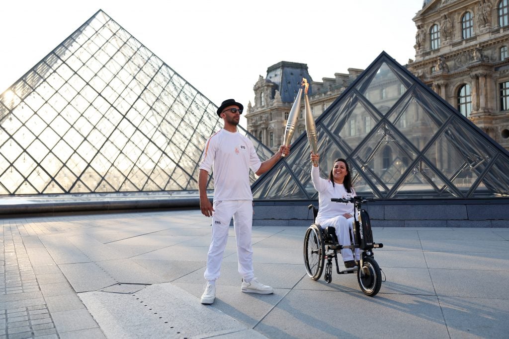 A man wearing sunglasses, a black fedora and a white shirt and top stands next to a dark haired woman in a wheelchair and matching white outfit. They are touching the tips of their silver Olympic torches together to pass the flame. They are in front of two large glass paneled pyramids in the courtyard of an ornate neoclassical building.