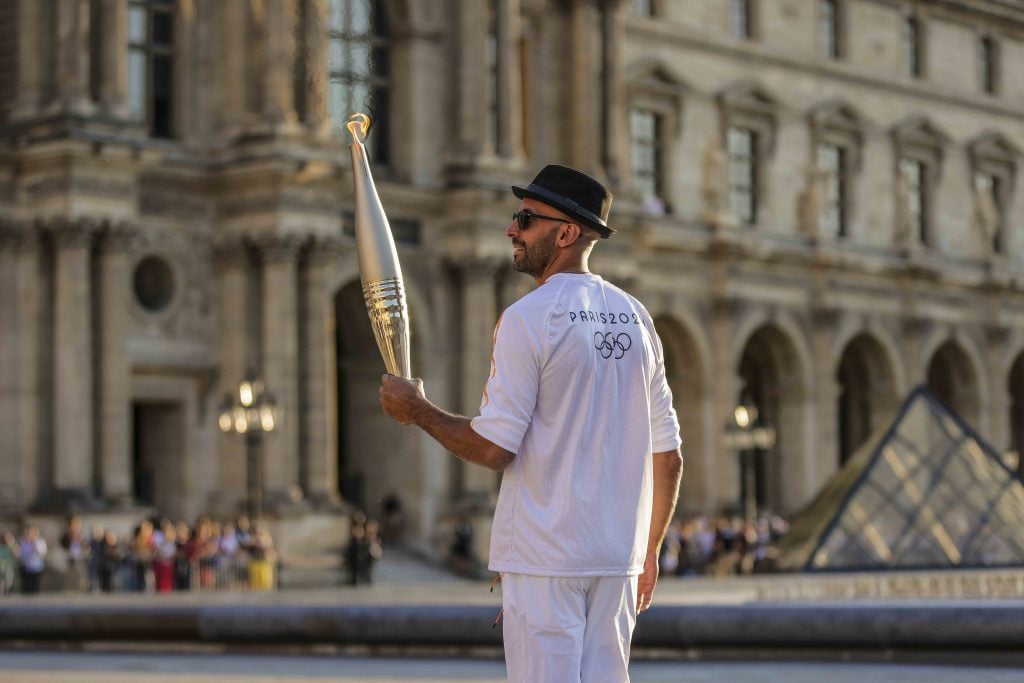 A man wearing sunglasses, a black fedora and a white shirt and top with a Paris Olympics 2024 logo on the back starts with his back to the camera, face turned to the side, holds up the silver Olympic torch, which is lit. He is standing in front of an ornate neoclassical building.