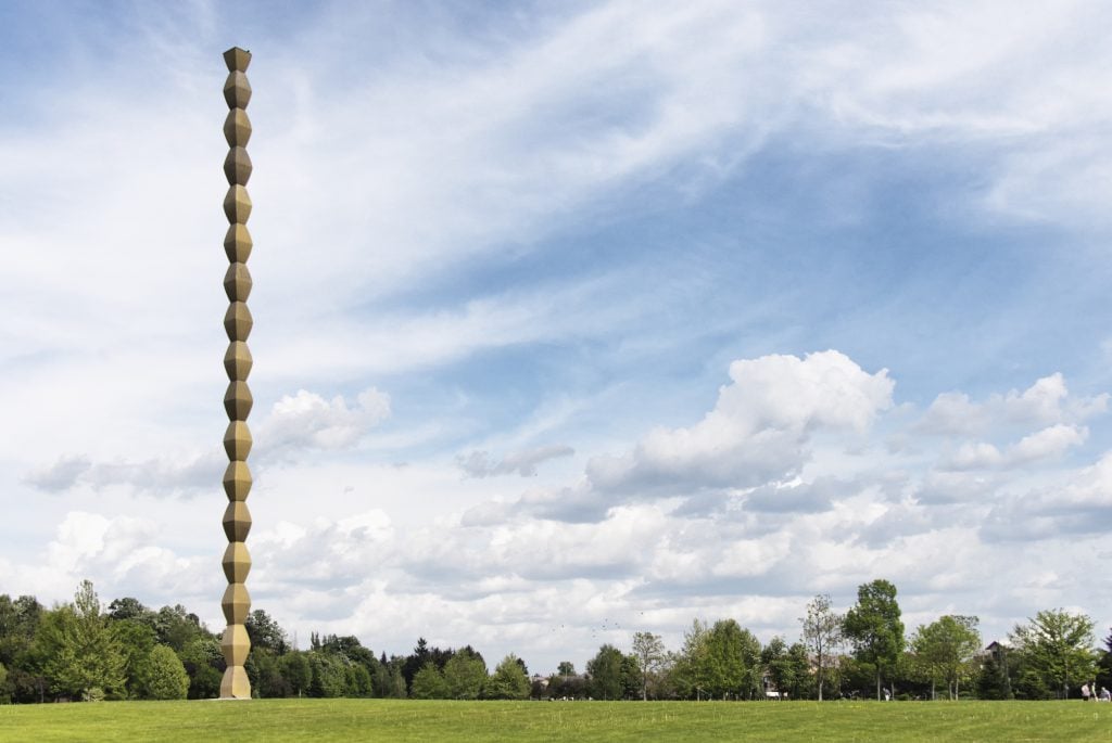a column made of 15 rhombus shapes rises into a sky with clouds above and a green field below