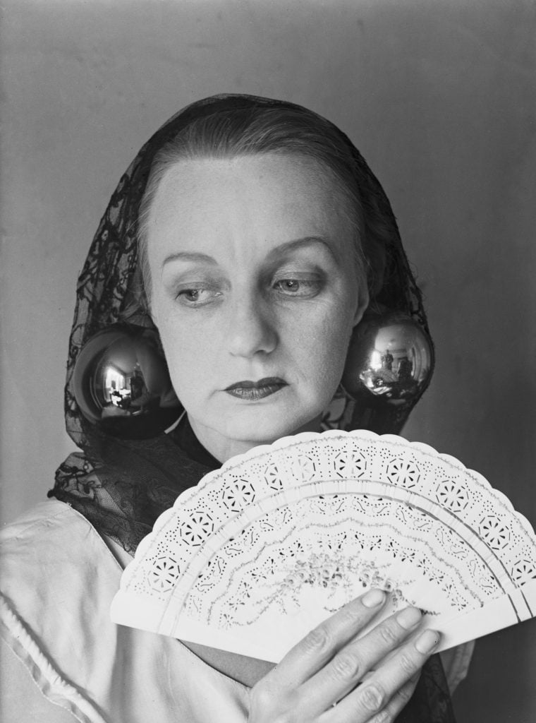 a black and white photo of a woman wearing large orb like earrings, a black mantilla and holding up a white fan