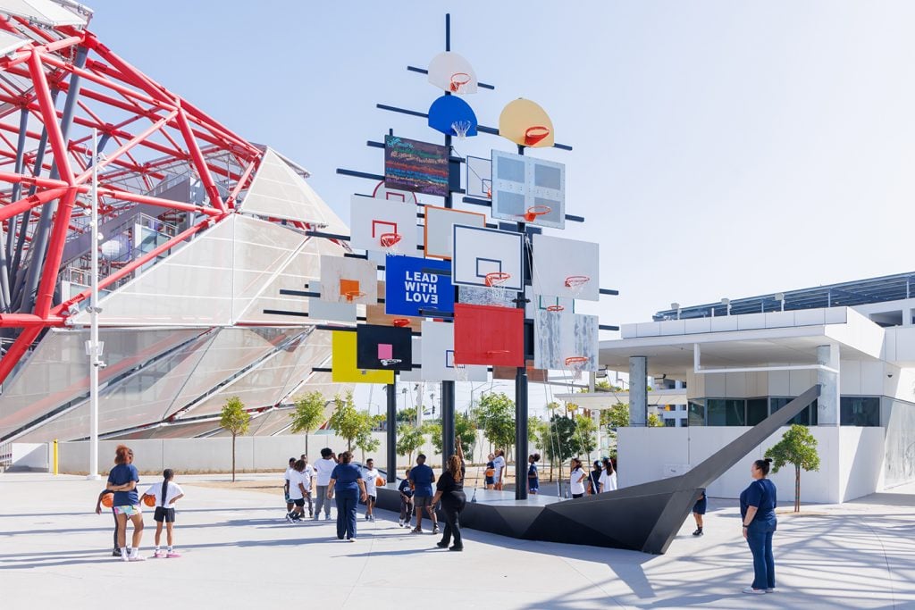 A sculptural installation featuring a cluster of basketball hoops arranged in a tree-like structure, with people, including children, interacting around it.