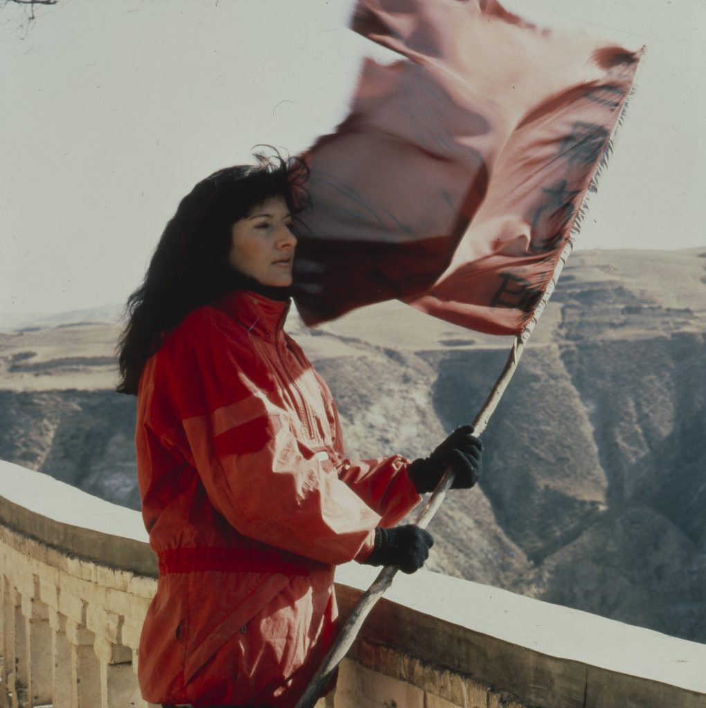 A young Marina Abramović in a red coat, holding a flag, atop the Great Wall of China for her 1988 performance piece with Ulay, "Great Wall Walk, China."