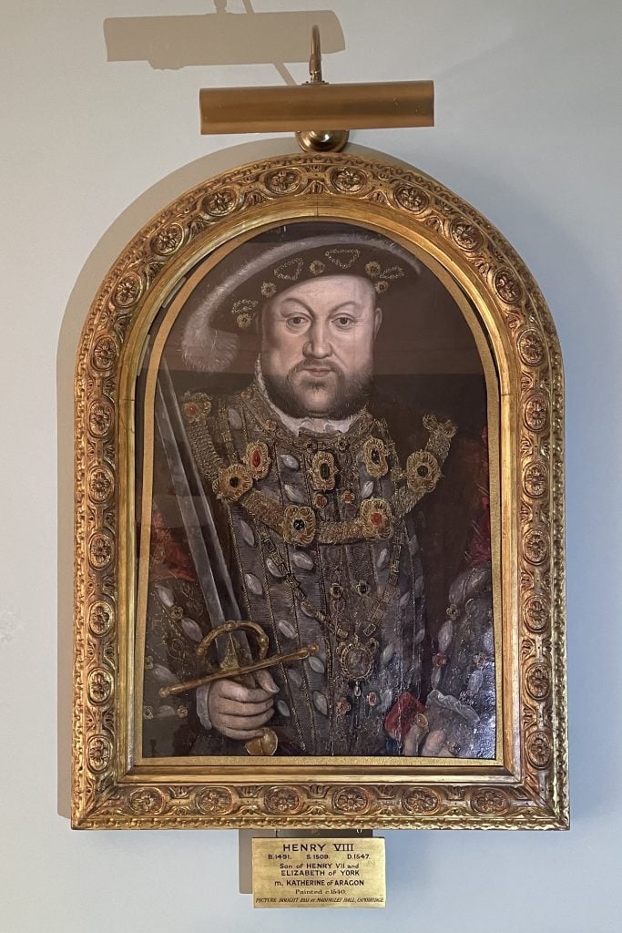 A framed portrait of Henry VIII displayed on a wall, featuring him in ornate royal attire with intricate jewelry and a feathered hat. He holds a sword, symbolizing his authority. The portrait is set within an elaborate gold frame with a plaque below that reads: "HENRY VIII B.1491 - S.1509 - D.1547 Son of HENRY VII and ELIZABETH OF YORK m. KATHERINE OF ARAGON Painted c. 1640." A picture light is positioned above the frame to illuminate the artwork.