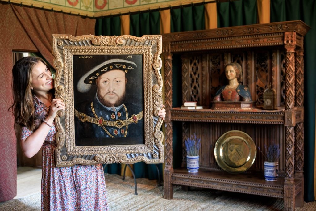 a woman holds up and smiles at an old fashioned painting of a man wearing a fancy hat, she is in a room that has walls draped in fabrics and a wooden cabinet