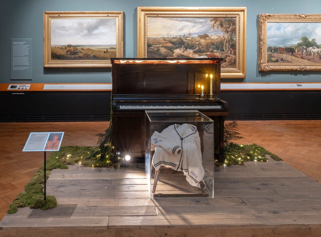 a cardigan with some moss near it on the floor and a white cardigan with a darker trim in a glass box, the room has old landscape paintings on the wall and blue walls