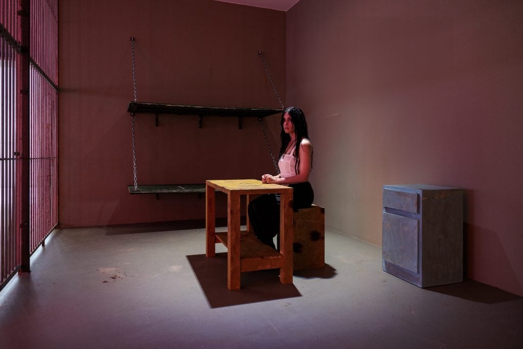 a woman sits at a desk in a sparsely furnished room with prison bars