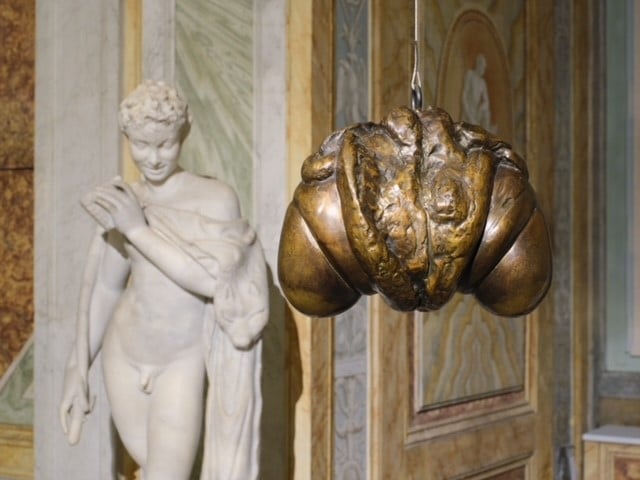 a bronze sculpture that vaguely resembles a croissant or a human crotch split open is hanging from a string in front of a classical marble nude statue