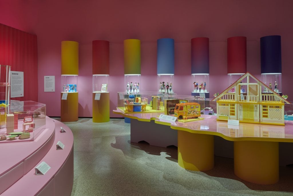 an exhibition gallery with pink walls, a green floor, and pastel shade pedestals featuring barbie dolls