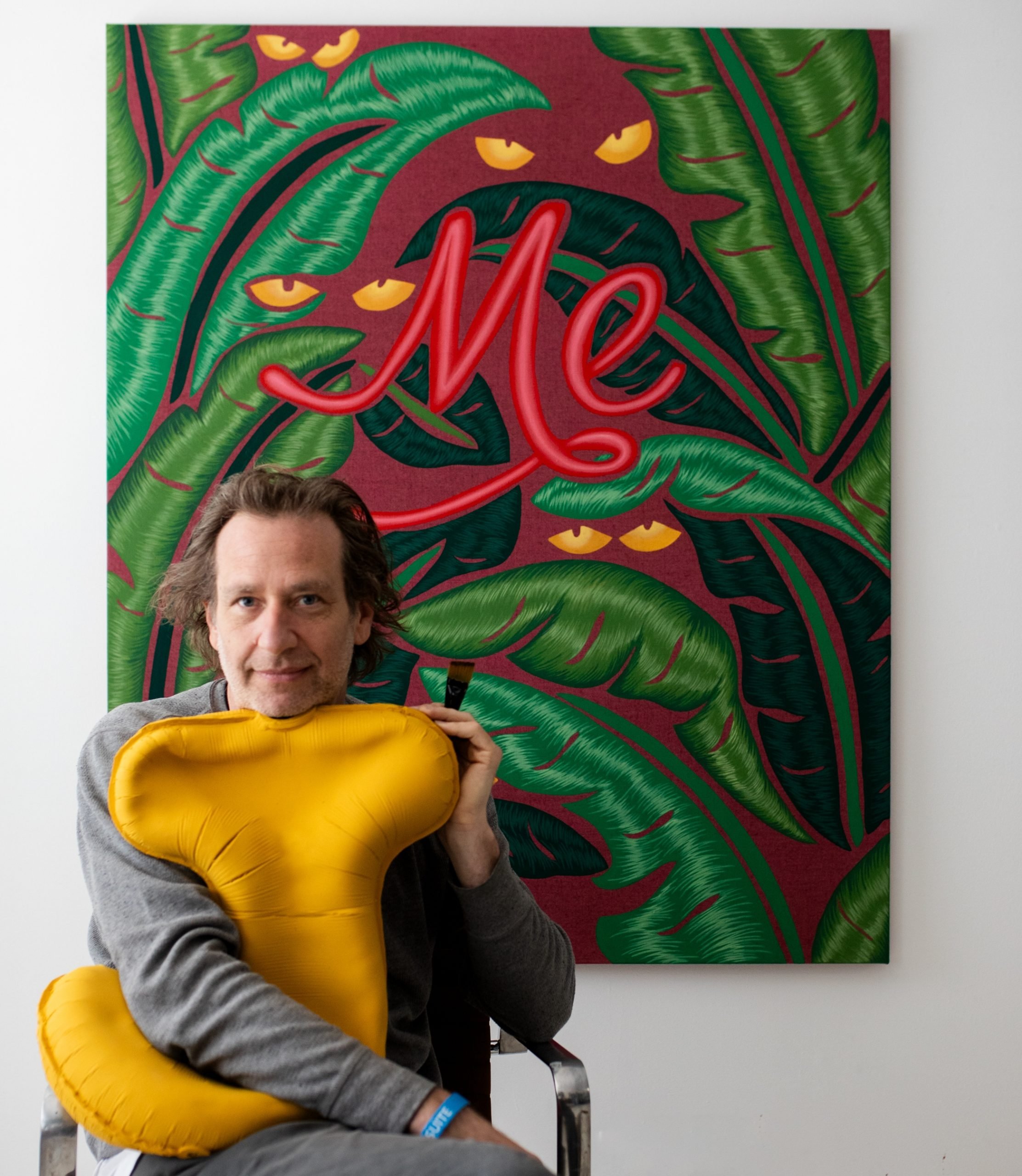 a man clutching a "J" shaped pillow sits in front of a painting with "me" scrawled in red letters and yellow eyes appearing to peer out of some shrubbery