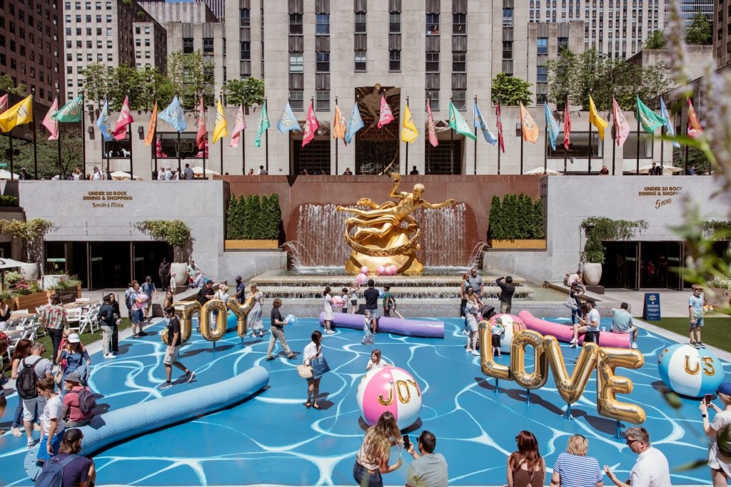 a replica of a pool against a gray inner city building, it has large blow up letters spelling love and large pool toys like a large beach ball saying "joy"