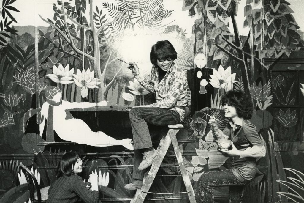 a smiling Kenzo Takada in the 1970s is flanked by two assistants and working on a vivid mural