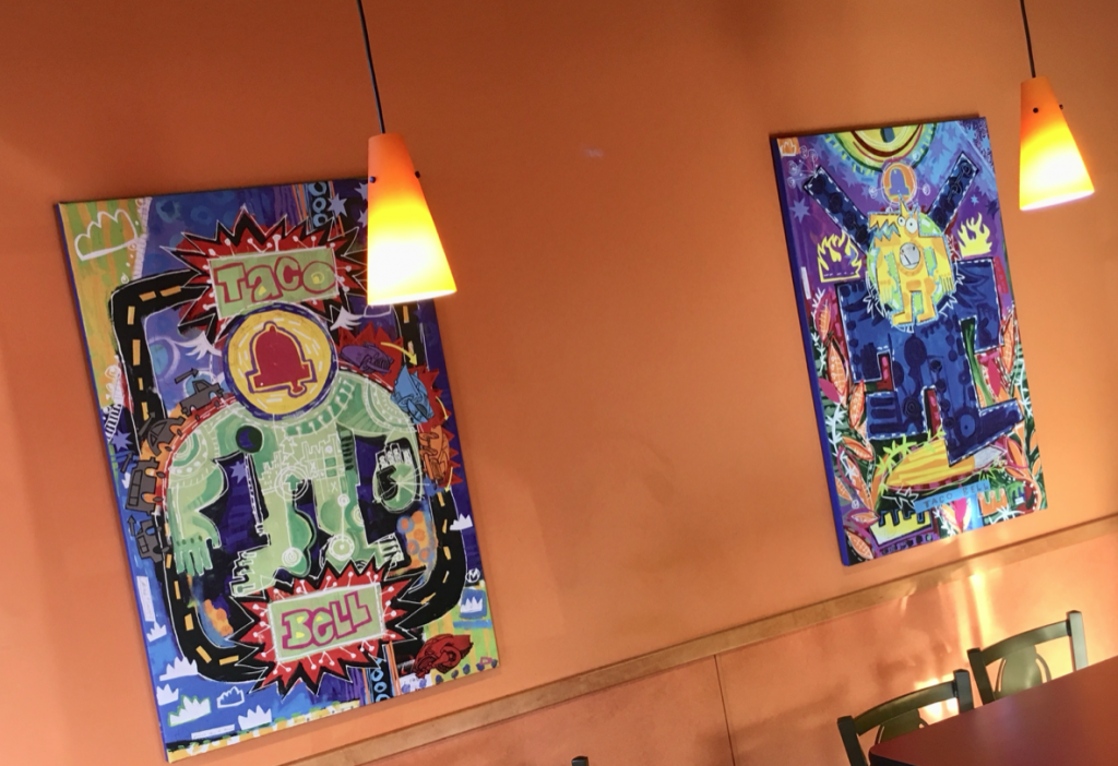 Two colorful paintings in a geometric style hanging on the orange wall of a Taco Bell. Two glowing light fixtures hang from the ceiling.