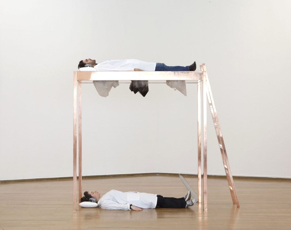 Marina Abramović's sculpture "Copper Bed for Human Use," made of copper and quartz stones, is seen in a gallery. The piece is a lofted bed with a ladder up to the top. One person is lying on the top, while another lies on the floor below beneath the three large crystals attached to the bottom of the bed frame. 