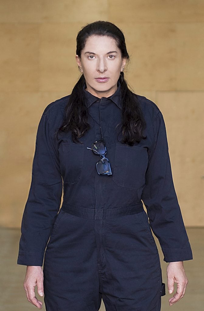Performance artist Marina Abramović clad in a navy blue jumpsuit. She has pale skin and long dark hair over both of her shoulders.
