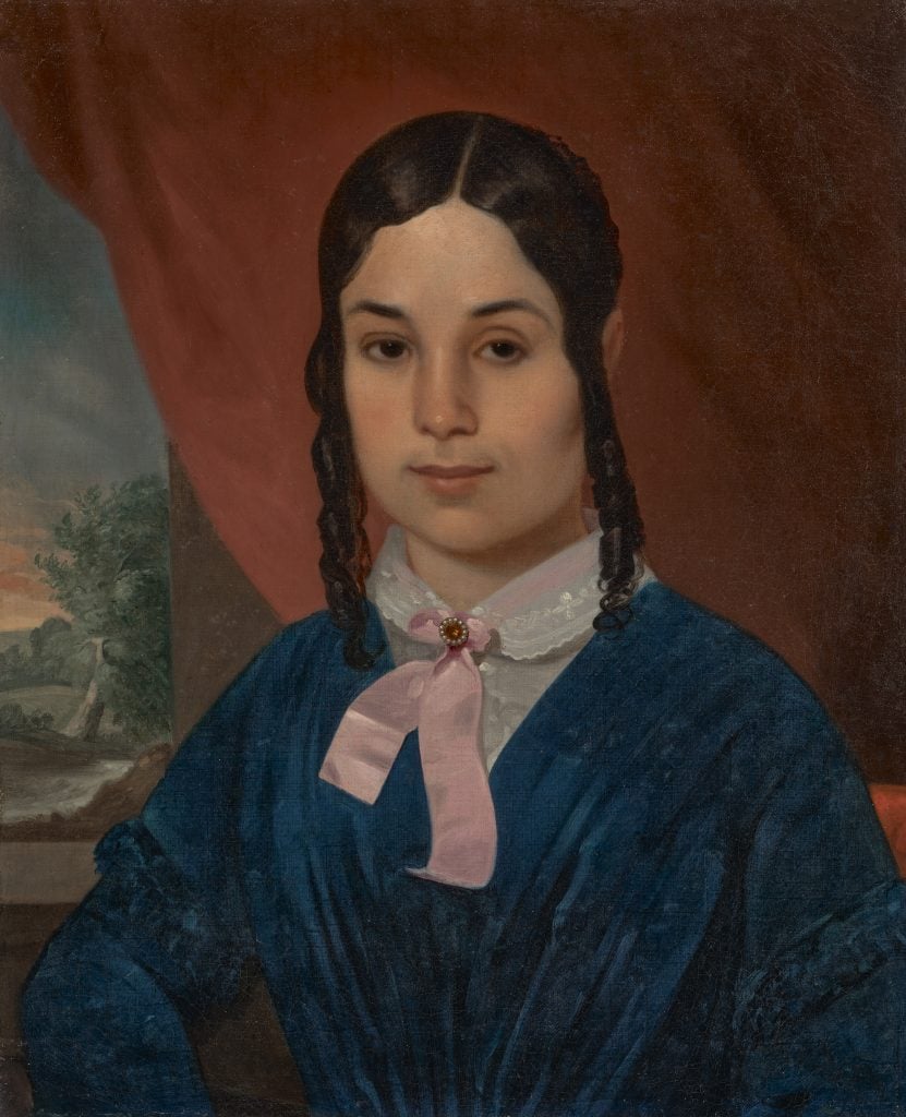 A 19th-century portrait of a young in a blue dress with a white color and a pink ribbon tied in a bow with a red brooch at the neck. Her dark brown hair is up with two long ringlets hanging on either side of her face. She is mixed race.