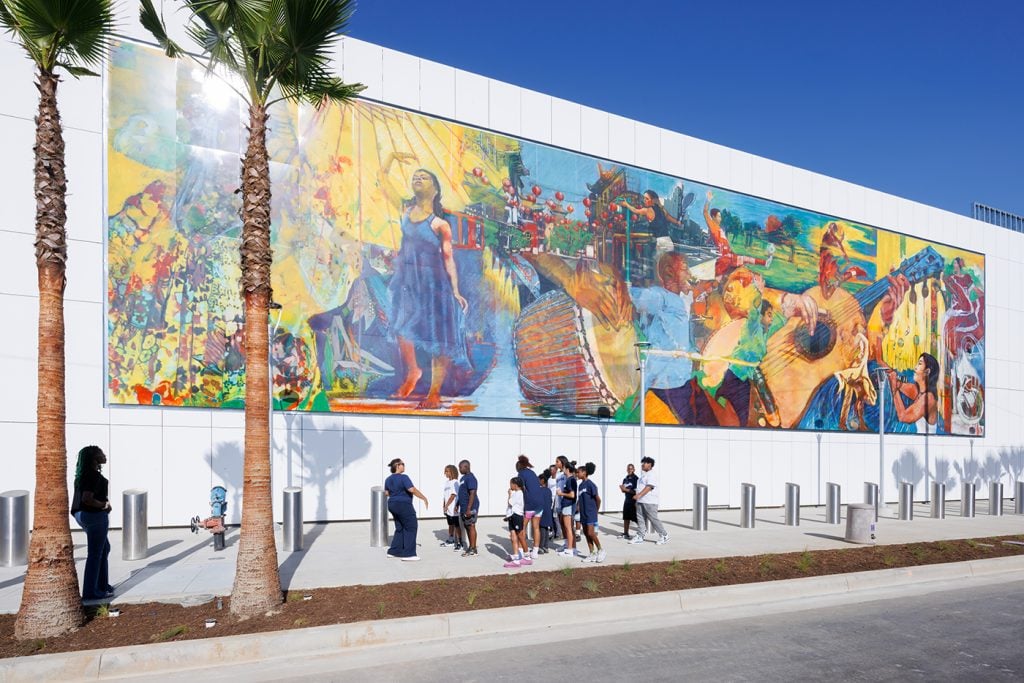 group of children and adults gather near a colorful, detailed mural on a white building, depicting diverse cultural scenes and activities.